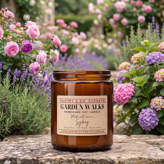 An amber glass jar candle labeled 'GARDEN WALKS' by Harvey & Co. Sydney sits on a stone surface outdoors, surrounded by blooming pink and purple flowers and green foliage.
