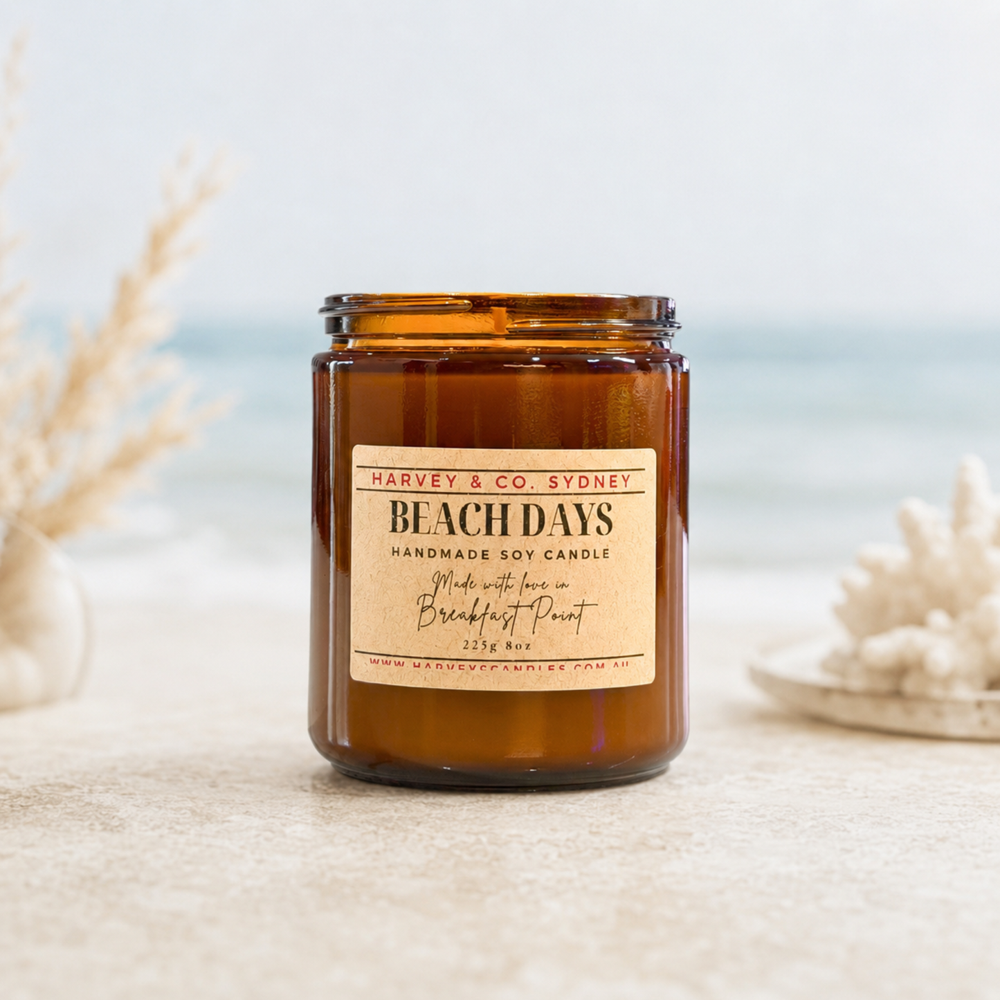 An amber glass jar candle labeled 'BEACH DAYS' by Harney & Co. Sydney, styled on a light stone surface with blurred seashells and a beach background.