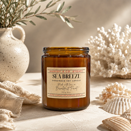 An amber glass jar candle labeled 'SEA BREEZE' by Harvey & Co. Sydney, styled on a beige surface with seashells, coral, a ceramic vase, and a folded linen cloth in a warm coastal-inspired setting.