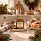 An amber glass jar candle labeled 'Cinnamon Scroll' sits on a kitchen counter surrounded by holiday decor, cinnamon sticks, and cinnamon rolls in a warm, softly lit kitchen setting.