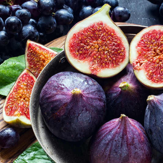 Fresh figs, including whole dark purple figs and several cut open to show pink-red interiors, arranged in a bowl with additional grapes and green leaves on a dark surface.