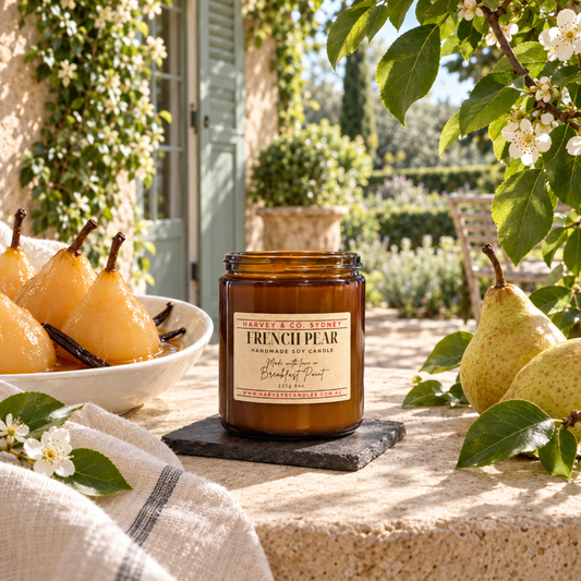 An amber glass jar candle labeled 'French Pear' sits on a stone coaster on a sunlit stone table outdoors, surrounded by pears, white flowers, green leaves, and a garden patio background.