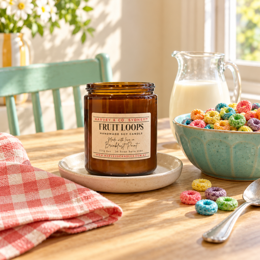 An amber glass jar candle labeled 'Fruit Loops' sits on a wooden table in a bright kitchen setting, surrounded by a bowl of colorful cereal, a pitcher of milk, a red-and-white checkered cloth, and a spoon.