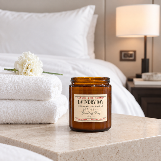 An amber glass jar candle labeled 'Harvey & Co. Sydney Laundry Day' sits on a light stone bedside table in a softly lit bedroom.