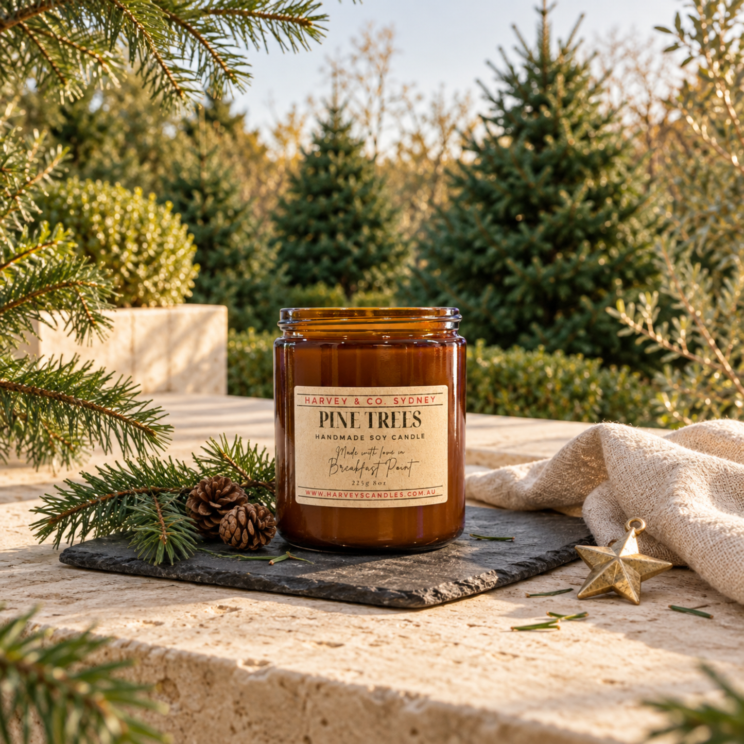 Harvey & Co. Sydney Pine Trees handmade soy candle in an amber glass jar, displayed on slate with pine branches, pinecones, and a gold star ornament outdoors among pine trees.