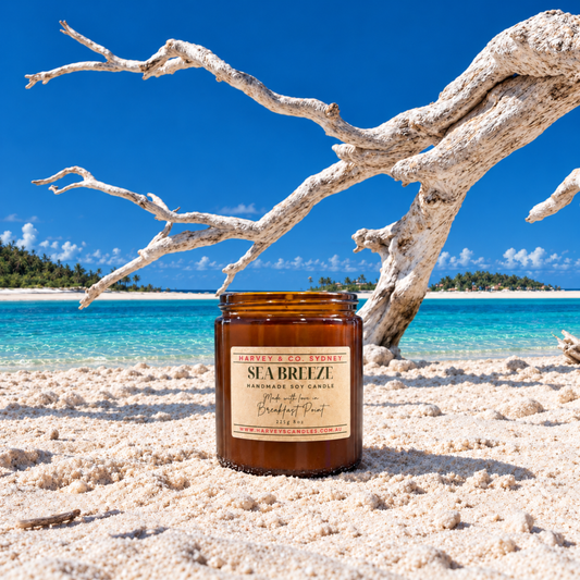 An amber glass jar candle labeled Sea Breeze by Beach and Vine stands on white sand in a tropical beach setting with turquoise water, a blue sky, and a large white driftwood branch behind it.