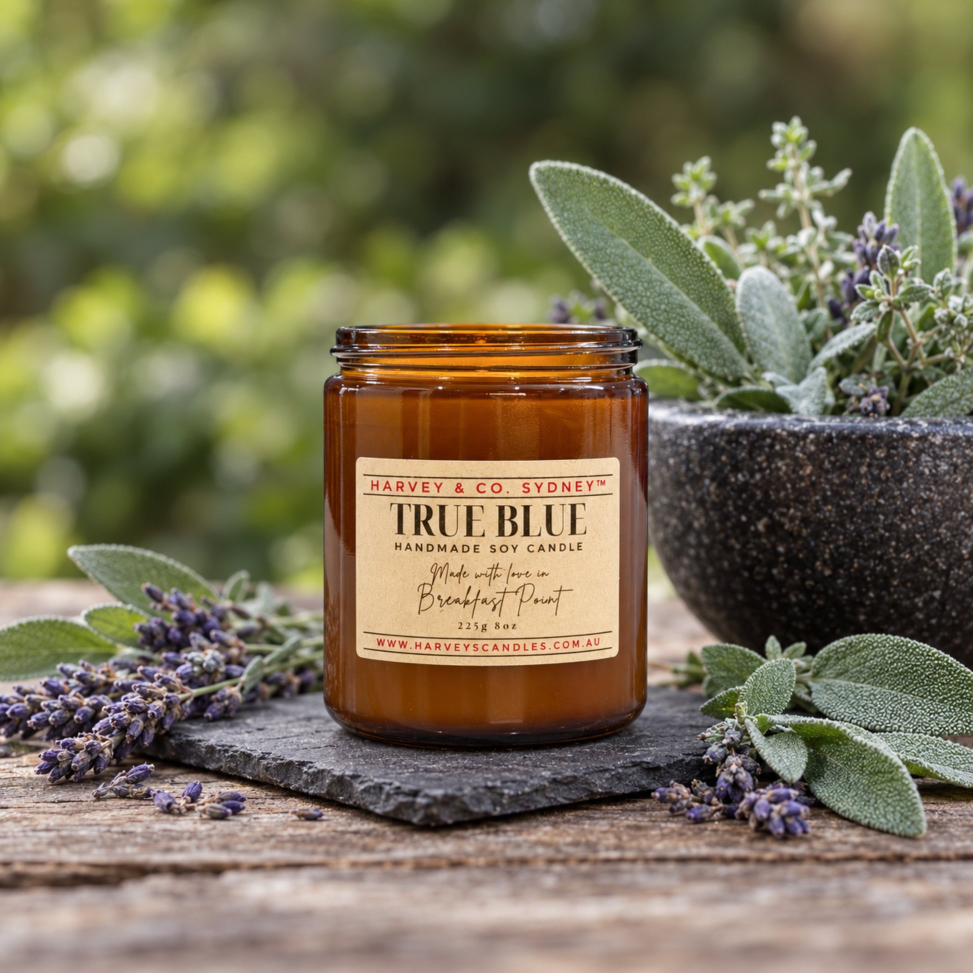 An amber glass jar candle labeled 'Harvest & Co. Sydney True Blue' sits on a rustic wooden surface with lavender sprigs, a stone plate, and a blurred garden background with green foliage.