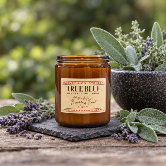 An amber glass jar candle labeled 'Harvest & Co. Sydney True Blue' sits on a rustic wooden surface with lavender sprigs, a stone plate, and a blurred garden background with green foliage.