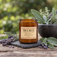 An amber glass jar candle labeled 'Harvest & Co. Sydney True Blue' sits on a rustic wooden surface with lavender sprigs, a stone plate, and a blurred garden background with green foliage.