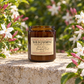 An amber glass jar candle labeled 'Wild Jasmine' by Harvey & Co. Sydney, placed on a stone ledge with blooming white and pink flowers in a sunlit garden background.