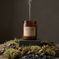 Amber glass soy candle labeled “Log Cabin” centered on a dark stone pedestal, surrounded by moss, bark, and dried natural elements on a forest-like surface. Thin smoke rises from the unlit wick against a moody brown background, creating a rustic, atmospheric studio scene.