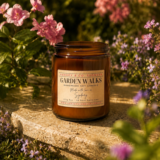 An amber glass jar candle labeled 'Garden Walks' sits on a stone surface outdoors in a garden with pink flowers and green foliage in soft sunlight.