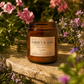An amber glass jar candle labeled 'Garden Walks' sits on a stone surface outdoors in a garden with pink flowers and green foliage in soft sunlight.
