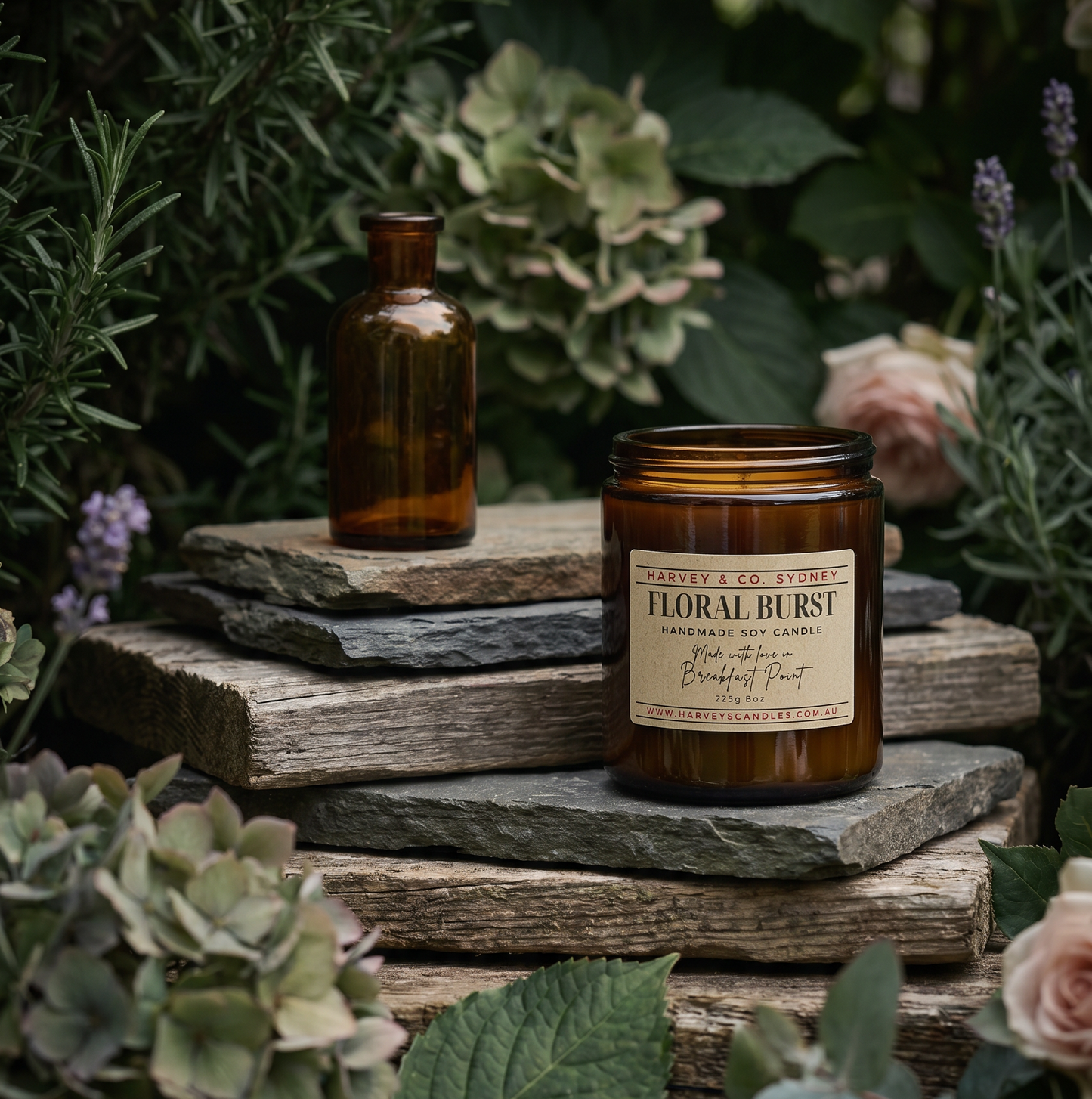 An amber glass jar candle labeled 'Floral Burst' sits on stacked rustic stones, with a matching empty amber bottle beside it. The scene is styled with green foliage, flowers, and wood in an outdoor garden-like setting.