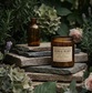 An amber glass jar candle labeled 'Floral Burst' sits on stacked rustic stones, with a matching empty amber bottle beside it. The scene is styled with green foliage, flowers, and wood in an outdoor garden-like setting.