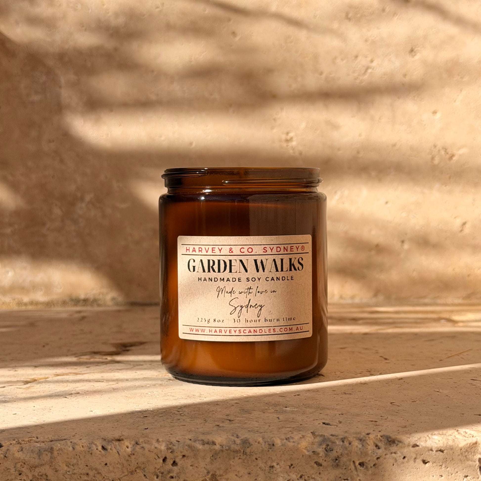 An amber glass jar candle labeled 'GARDEN WALKS' by Harvey & Co. Sydney, placed on a stone surface with warm sunlight and leaf shadows across the scene.