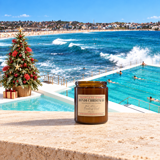 A jar candle labeled 'Bondi Christmas' sits on a stone ledge overlooking Bondi Beach and the ocean, with a decorated Christmas tree and beachgoers in the pool in the background.