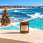 A jar candle labeled 'Bondi Christmas' sits on a stone ledge overlooking Bondi Beach and the ocean, with a decorated Christmas tree and beachgoers in the pool in the background.