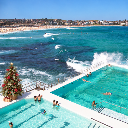 A high-angle outdoor view of a coastal beach and ocean with two large swimming pools in the foreground, people swimming and relaxing, a sandy beach, buildings along the shoreline, and a decorated Christmas tree near the pool.