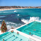 A high-angle outdoor view of a coastal beach and ocean with two large swimming pools in the foreground, people swimming and relaxing, a sandy beach, buildings along the shoreline, and a decorated Christmas tree near the pool.