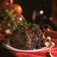 Christmas gourmand scented candle in amber jar with holly leaves and a lit wick on a festive table.