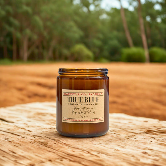 Brown candle labeled 'True Blue' on a wooden surface with a blurred natural background