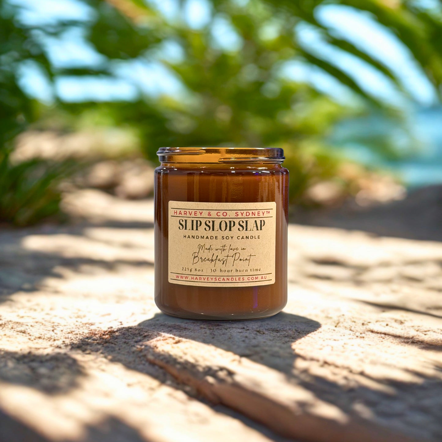 Harvey & Co. Sydney candle labeled 'Slip SlipSlap' on a sandy surface with blurred greenery in the background