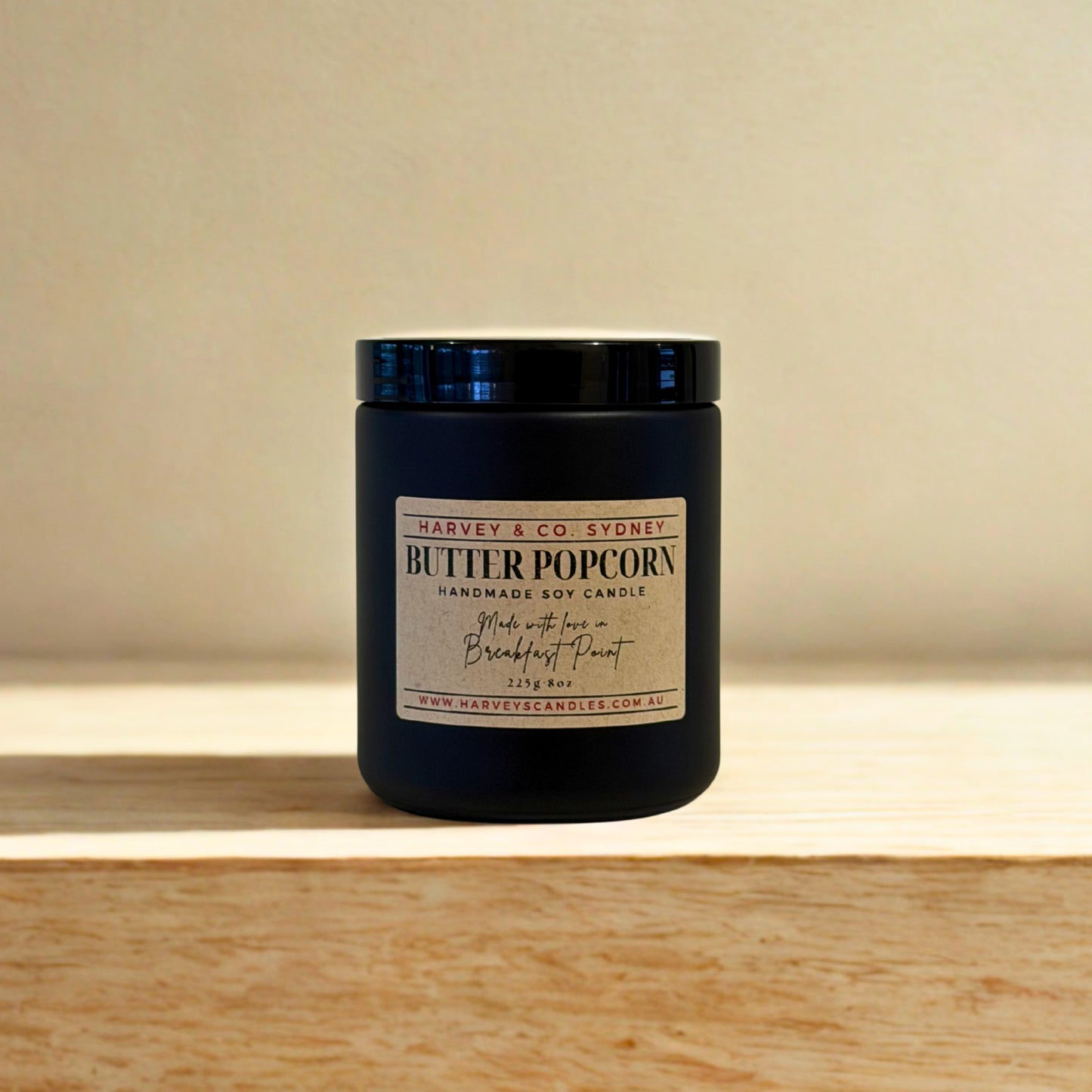 Black candle labeled 'Butter Popcorn' by Harvey & Co. Sydney on a wooden surface with a beige background.