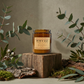 Candle labeled 'Bush Walks' on a wooden block with eucalyptus leaves around