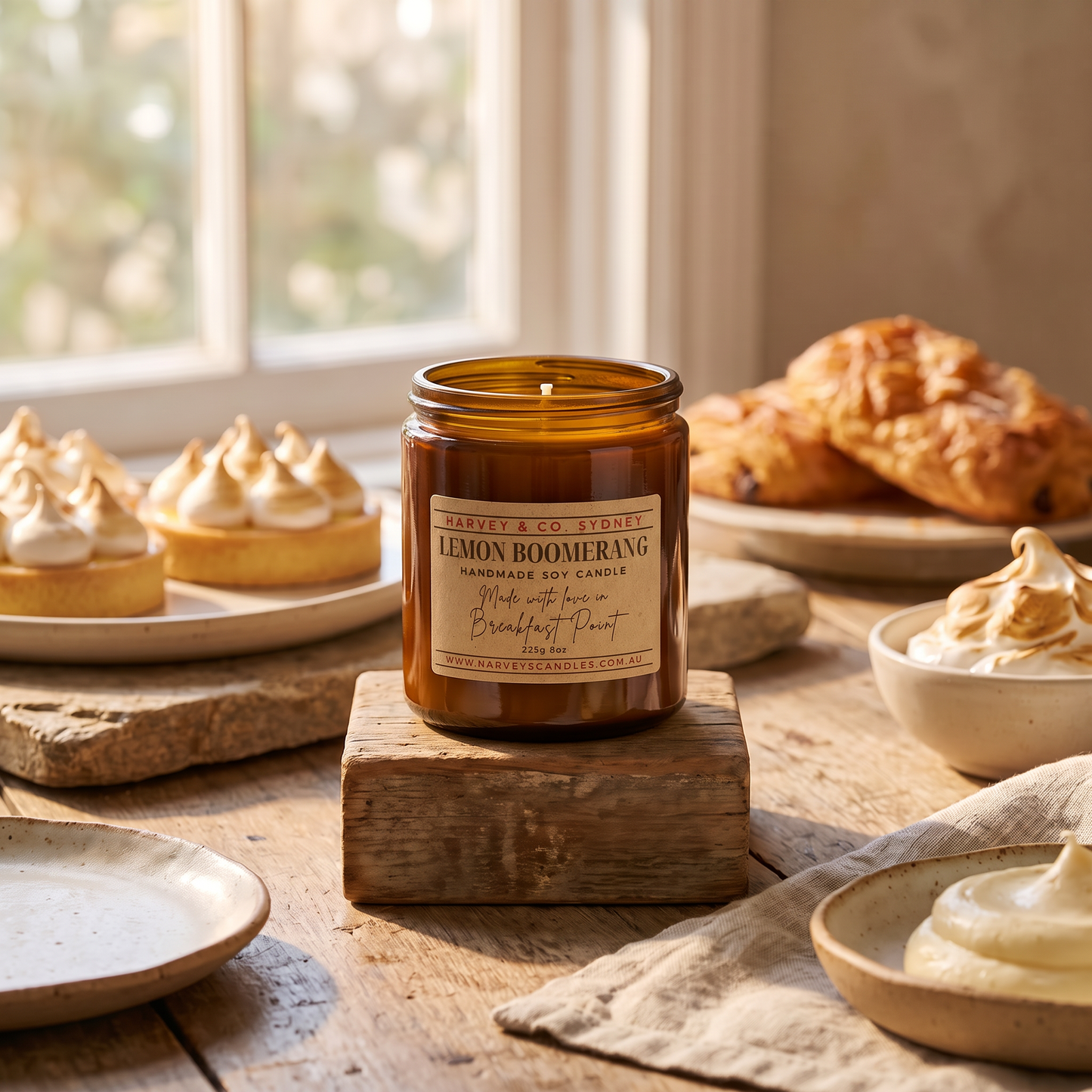 An amber glass candle jar labeled 'Lemon Boomerang' by Saviers Co. London, displayed on a rustic wooden block on a table with pastries and cream in the background.