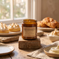 An amber glass candle jar labeled 'Lemon Boomerang' by Saviers Co. London, displayed on a rustic wooden block on a table with pastries and cream in the background.