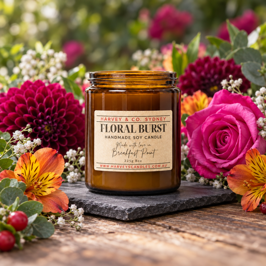 An amber glass jar candle labeled 'Harvey & Co Sydney Floral Burst' sits on a dark slate tray surrounded by large pink and orange flowers on a rustic wooden surface with a blurred garden background.