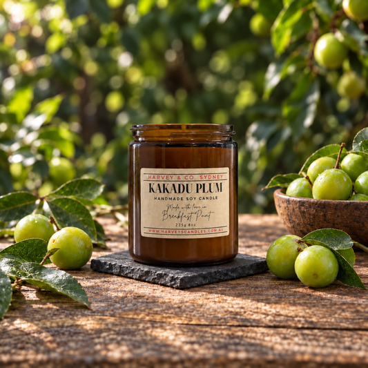 An amber glass jar candle labeled 'KAKADU PLUM' sits on a dark stone coaster on a rustic wooden table, surrounded by fresh green plums and leaves in an outdoor orchard setting.
