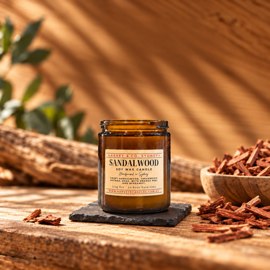 An amber glass jar candle labeled Sandalwood Background Scents sits on a dark slate coaster on a rustic wooden table, surrounded by wood chips and a bowl, with a warm wood wall and leaf shadows in the background.