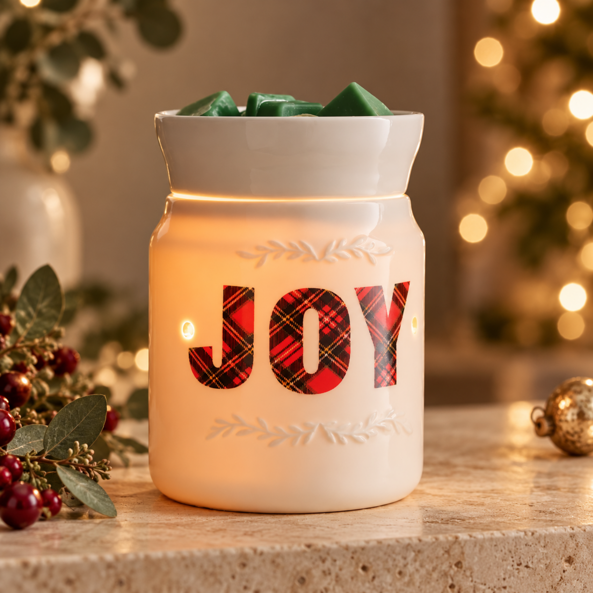 A white ceramic Christmas wax warmer candle with the word 'JOY' in red plaid letters, lit with warm internal lights, set on a stone surface with holiday decorations and blurred Christmas lights in the background.