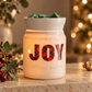 A white ceramic Christmas wax warmer candle with the word 'JOY' in red plaid letters, lit with warm internal lights, set on a stone surface with holiday decorations and blurred Christmas lights in the background.